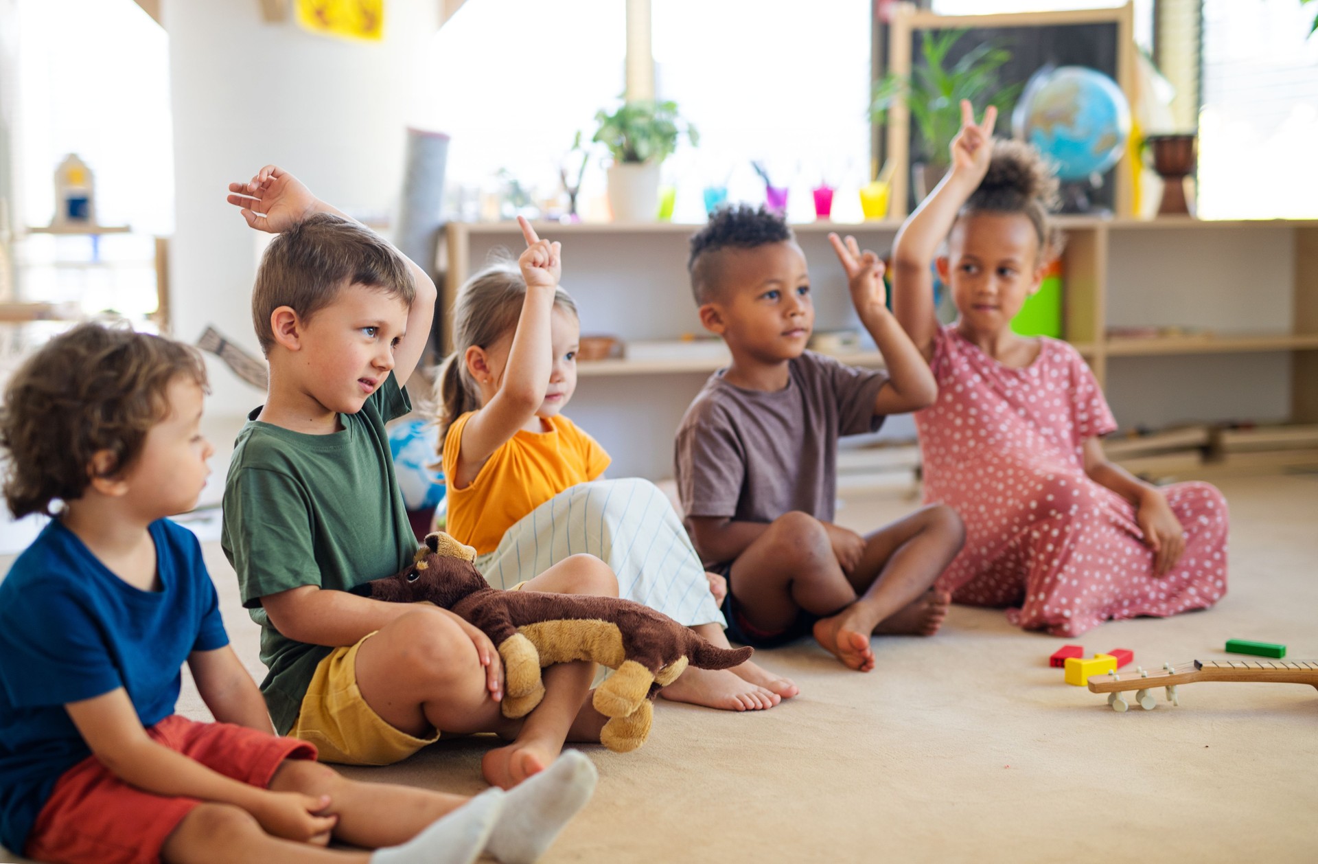 Group of small nursery school children sitting on floor indoors in classroom, raising hands.